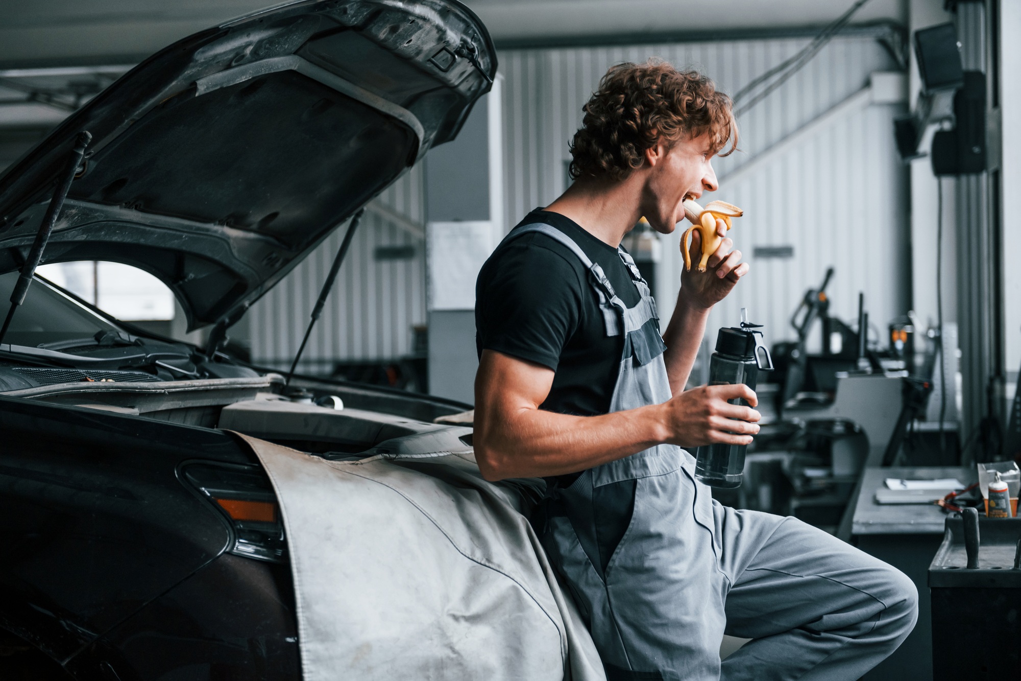 Eats banana and drinks water. Adult man in grey colored uniform works in the automobile salon